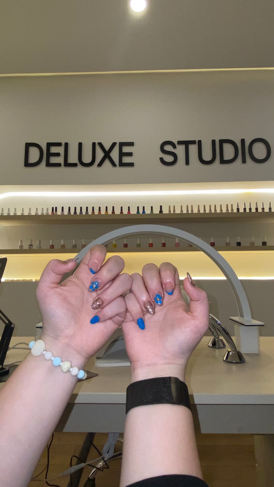 Manicure en azul eléctrico con detalles dorados, frente a la estantería de esmaltes